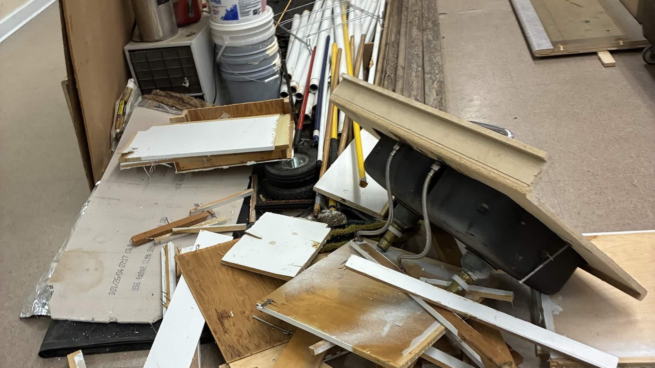 Stripped cabinet doors, trim boards, plywood panels and a cast-iron sink piled along a corridor wall, with buckets and tools stacked behind them.